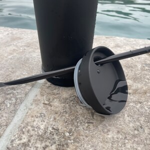 May include: A black travel mug with a black lid and a black straw. The mug is sitting on a grey tiled surface.