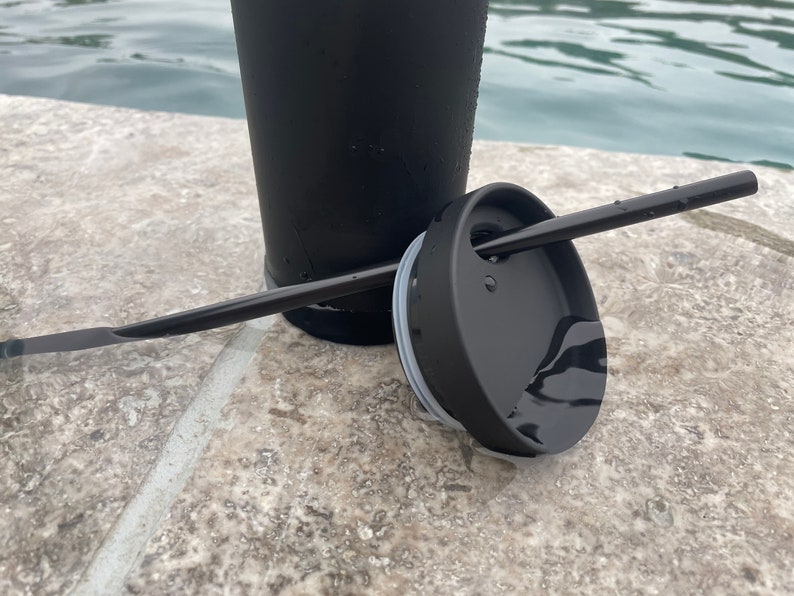 May include: A black travel mug with a black lid and a black straw. The mug is sitting on a grey tiled surface.