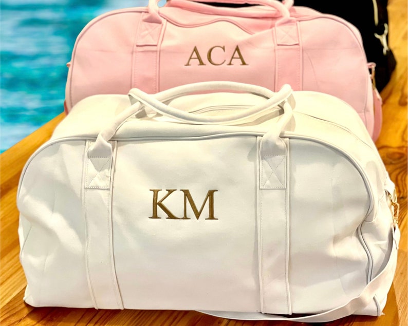 May include: Two white canvas duffel bags with gold embroidered initials. The bag on top has the initials "ACA" and the bag on the bottom has the initials "KM".