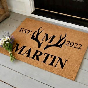 May include: A rectangular doormat made of natural coir fiber. The mat features the text "EST. MARTIN 2022" in black, with stylized antlers and the letter "M" in the center. A small bouquet of flowers rests on the left side.