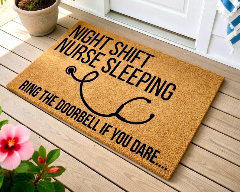 May include: A rectangular doormat with the text "NIGHT SHIFT NURSE SLEEPING RING THE DOORBELL IF YOU DARE..." in black. A black stethoscope graphic is centered on the mat. The mat is a light brown color and is placed on a wooden porch.