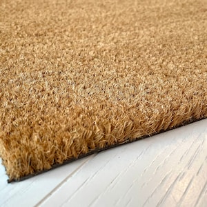 May include: Close-up of a rectangular doormat made of natural coir fibers. The mat has a light brown color and a textured surface. The edges are dark brown, and the mat is placed on a white surface.