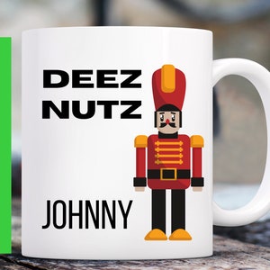 May include: White ceramic mug with a red and yellow cartoon nutcracker holding a rifle. The mug has the text "DEEZ NUTZ" and "JOHNNY" printed on it.