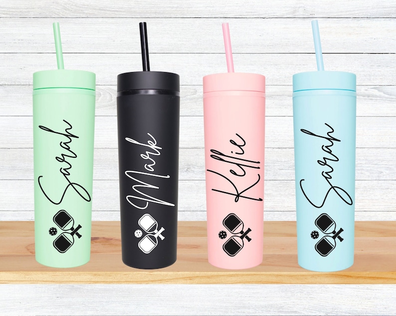 K&ouml;nnte beinhalten: Vier personalisierte hohe Becher mit Strohhalmen. Die Becher sind in Gr&uuml;n, Schwarz, Rosa und Blau. Jeder Becher hat einen Namen darauf geschrieben: Sarah, Mark, Kellie und Sarah. Jeder Becher hat ein Schwarzwei&szlig;bild von zwei Paddleball-Schl&auml;gern und einem Ball.