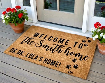 May include: A rectangular doormat made of natural coir fiber. The mat features the text "WELCOME TO The Smith home OK, OK.. LOLA'S HOME!" in black script. Black paw prints are scattered across the mat.
