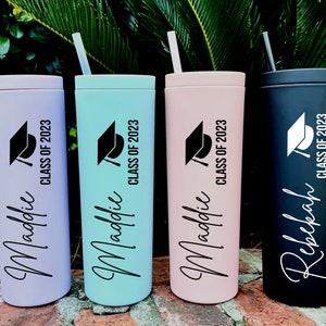 Puede incluir: Cuatro vasos de graduaci&oacute;n personalizados con pajitas. Los vasos son de color lavanda, verde azulado, rosa y negro. Cada vaso tiene un gorro de graduaci&oacute;n negro y el texto "Clase de 2023". Los nombres "Maddie" y "Rebekah" est&aacute;n escritos en los vasos.