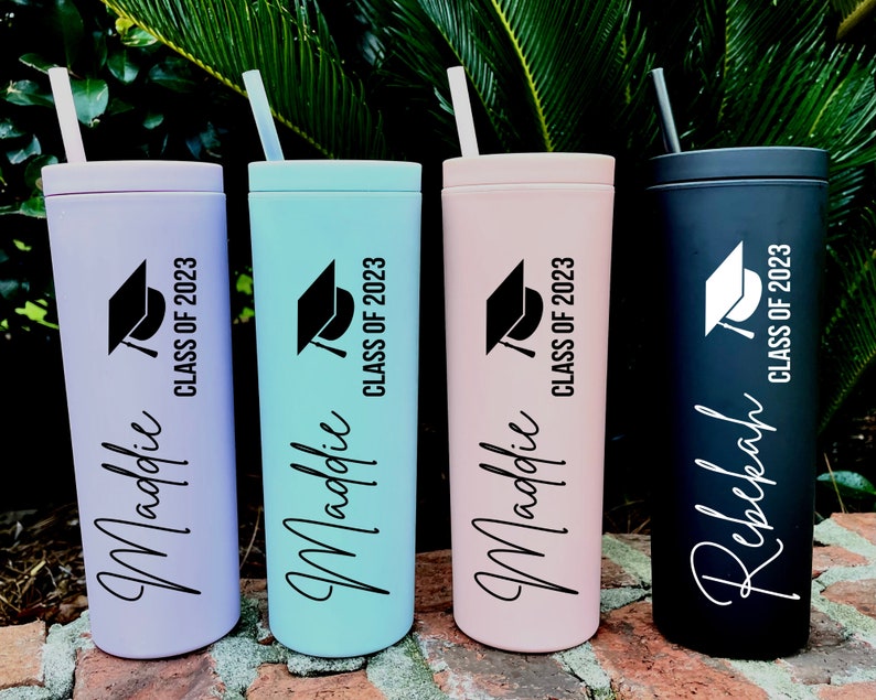 Puede incluir: Cuatro vasos de graduaci&oacute;n personalizados con pajitas. Los vasos son de color lavanda, verde azulado, rosa y negro. Cada vaso tiene un gorro de graduaci&oacute;n negro y el texto "Clase de 2023". Los nombres "Maddie" y "Rebekah" est&aacute;n escritos en los vasos.