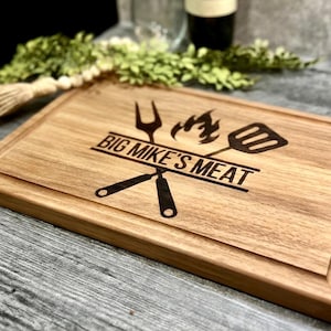 Puede incluir: Tabla de cortar rectangular de madera clara con ranura para jugos. La tabla presenta el texto "BIG MIKE'S MEAT" con un tenedor, una esp&aacute;tula y una llama.