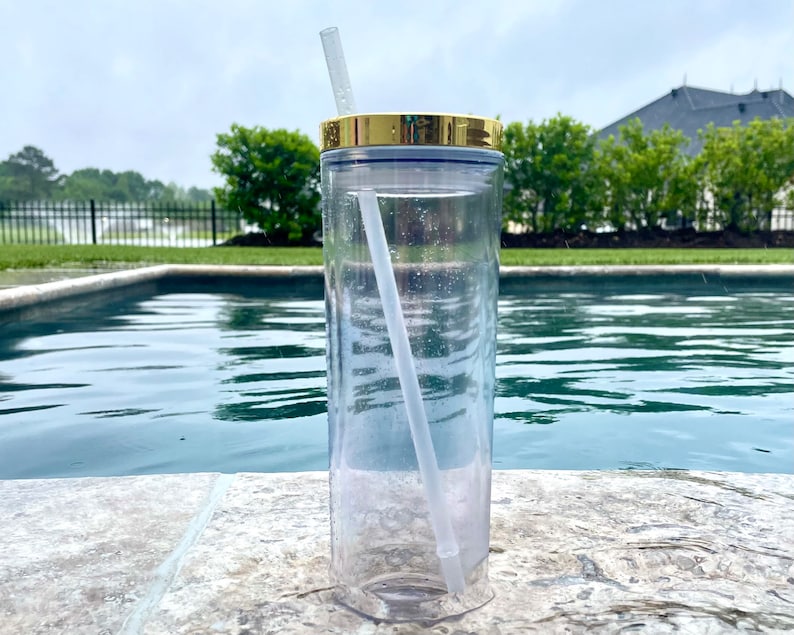 May include: A clear plastic tumbler with a gold lid and a clear plastic straw. The tumbler is sitting on a concrete surface next to a swimming pool.