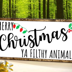 May include: A white wooden sign with a brown frame. The sign says "Merry Christmas Ya Filthy Animal!" in black text. There are red and green Christmas lights and a green holly sprig with red berries on the sign.