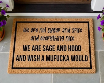 May include: A rectangular doormat with a black border and text that reads "We are not sugar and spice and everything nice WE ARE SAGE AND HOOD AND WISH A MUFUCKA WOULD" in black lettering. The mat is a light brown color.