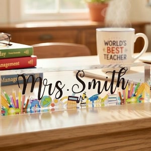 May include: A clear acrylic desk nameplate with the name "Mrs. Smith" in black script. The nameplate is decorated with colorful school-themed illustrations, including pencils, a globe, and books. A mug that says "World's Best Teacher" is in the background.