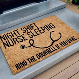 May include: A rectangular doormat made of natural coir fiber. The mat features black text that reads "NIGHT SHIFT NURSE SLEEPING" with a stethoscope graphic and "RING THE DOORBELL IF YOU DARE...."