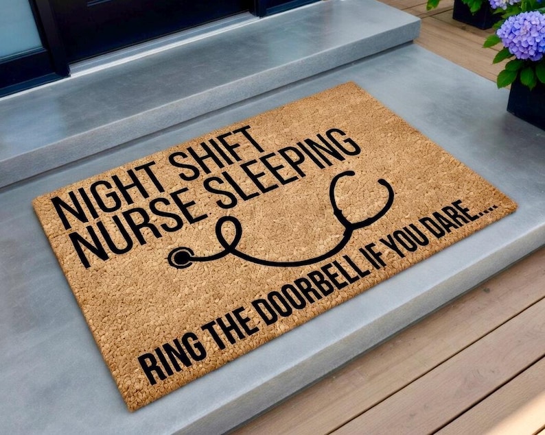 May include: A rectangular doormat made of natural coir fiber. The mat features black text that reads "NIGHT SHIFT NURSE SLEEPING" with a stethoscope graphic and "RING THE DOORBELL IF YOU DARE...."