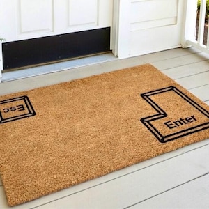 May include: A rectangular doormat made of natural brown coir fiber. The mat features a black outline of an "Enter" key and an "Esc" key, with the words "Enter" and "Esc" printed in black. The mat is placed on a wooden porch.