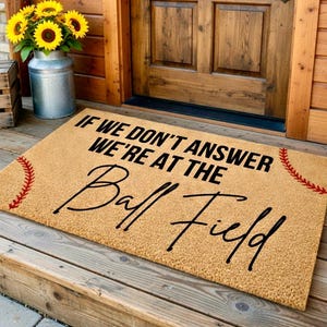 Puede incluir: Felpudo de fibra de coco beige con el texto "IF WE DON'T ANSWER WE'RE AT THE Ball Field" en negro. Costuras rojas de b&eacute;isbol adornan las esquinas. La alfombra est&aacute; en un porche de madera.