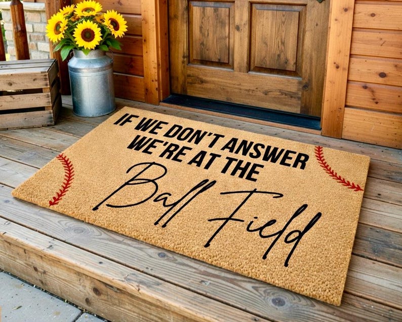 Puede incluir: Felpudo de fibra de coco beige con el texto "IF WE DON'T ANSWER WE'RE AT THE Ball Field" en negro. Costuras rojas de b&eacute;isbol adornan las esquinas. La alfombra est&aacute; en un porche de madera.