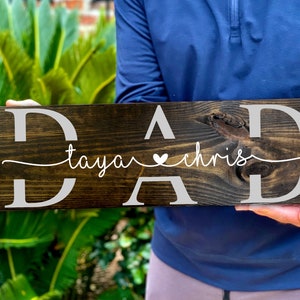 May include: A dark brown wooden sign with the word "DAD" in white letters. The sign also has the names "taya" and "chris" written in white cursive with a small heart symbol between them.