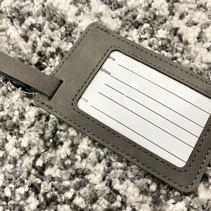 Peut inclure: Etiquette &agrave; bagages en cuir gris avec une &eacute;tiquette blanche et des bords cousus. L'&eacute;tiquette comporte des lignes pour &eacute;crire un nom, une adresse et un num&eacute;ro de t&eacute;l&eacute;phone.