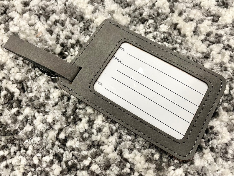 Peut inclure: Etiquette &agrave; bagages en cuir gris avec une &eacute;tiquette blanche et des bords cousus. L'&eacute;tiquette comporte des lignes pour &eacute;crire un nom, une adresse et un num&eacute;ro de t&eacute;l&eacute;phone.