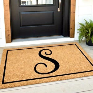 May include: A rectangular tan coir doormat featuring a black border and a large, centered black letter S in a cursive script font. The mat is positioned on a light-colored wooden porch in front of a black door with a green potted plant nearby.