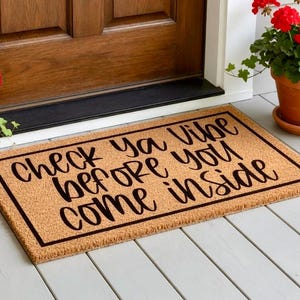 May include: A rectangular coir doormat with the text "Check ya vibe before you come inside" in black script. The mat has a black border and is placed in front of a wooden door. Potted red flowers are on either side.