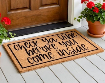 May include: A rectangular coir doormat with the text "Check ya vibe before you come inside" in black script. The mat has a black border and is placed in front of a wooden door. Potted red flowers are on either side.
