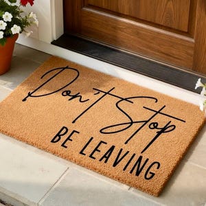 Peut inclure: Un paillasson rectangulaire en fibre de coco avec les mots "Don't Stop BE LEAVING" en écriture noire. Le tapis est beige et se trouve devant une porte en bois. Des plantes en pot avec des fleurs rouges et blanches sont de chaque côté.