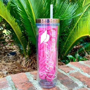 Puede incluir: Un vaso transparente de pl&aacute;stico con una pajita rosa y una tapa dorada. El vaso tiene un dise&ntilde;o blanco que dice "Jessica Class of 2022" y un gr&aacute;fico de un gorro de graduaci&oacute;n.