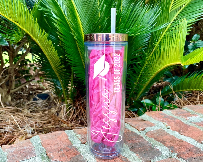 Puede incluir: Un vaso transparente de pl&aacute;stico con una pajita rosa y una tapa dorada. El vaso tiene un dise&ntilde;o blanco que dice "Jessica Class of 2022" y un gr&aacute;fico de un gorro de graduaci&oacute;n.