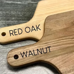 Puede incluir: Dos tablas de cortar de madera, una clara y otra oscura, con asas. La tabla clara lleva la inscripci&oacute;n "RED OAK" y la tabla oscura lleva la inscripci&oacute;n "WALNUT" en texto negro. Ambas tablas tienen un agujero en el asa para colgar.