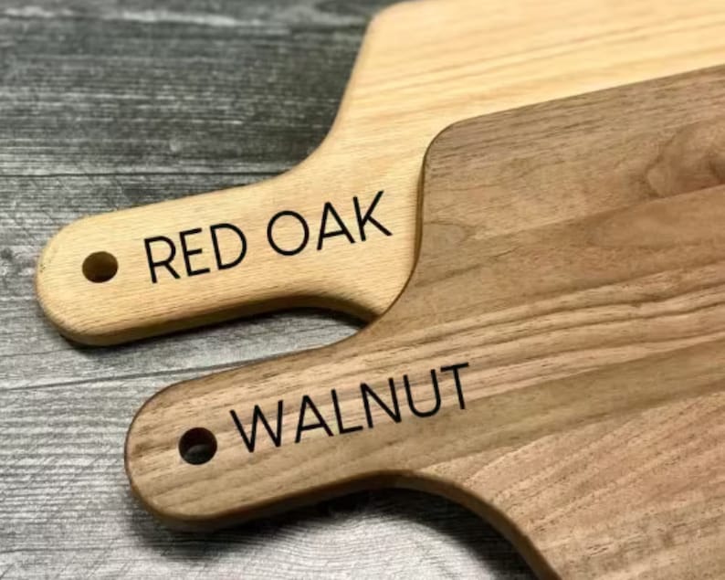 Puede incluir: Dos tablas de cortar de madera, una clara y otra oscura, con asas. La tabla clara lleva la inscripci&oacute;n "RED OAK" y la tabla oscura lleva la inscripci&oacute;n "WALNUT" en texto negro. Ambas tablas tienen un agujero en el asa para colgar.