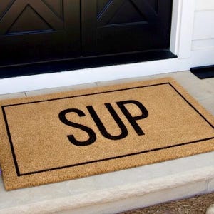 May include: A rectangular doormat made of natural coir fiber. The mat has a brown background with a black border and the word "SUP" in large, bold, black letters. The mat is placed on a concrete step in front of a black door.