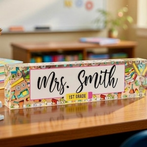 May include: A clear acrylic desk nameplate with the name "Mrs. Smith" in black script, and "1st Grade" in a smaller font. The nameplate is decorated with colorful school-themed illustrations.