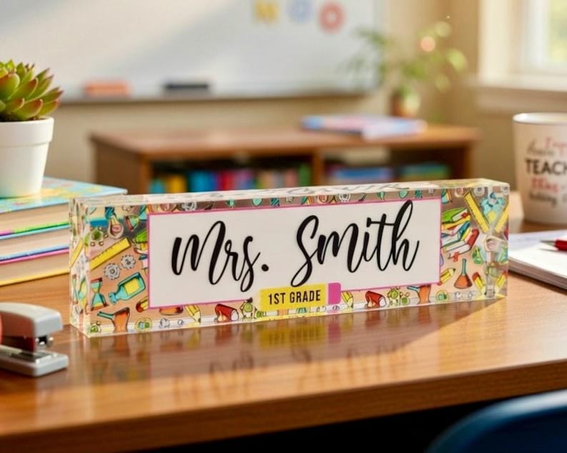 May include: A clear acrylic desk nameplate with the name "Mrs. Smith" in black script, and "1st Grade" in a smaller font. The nameplate is decorated with colorful school-themed illustrations.