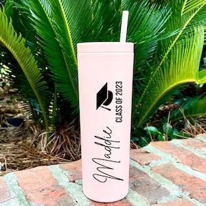 Puede incluir: Un vaso de acero inoxidable rosa con un gorro de graduaci&oacute;n negro y el texto "Class of 2023" y "Maddie" en letras negras.