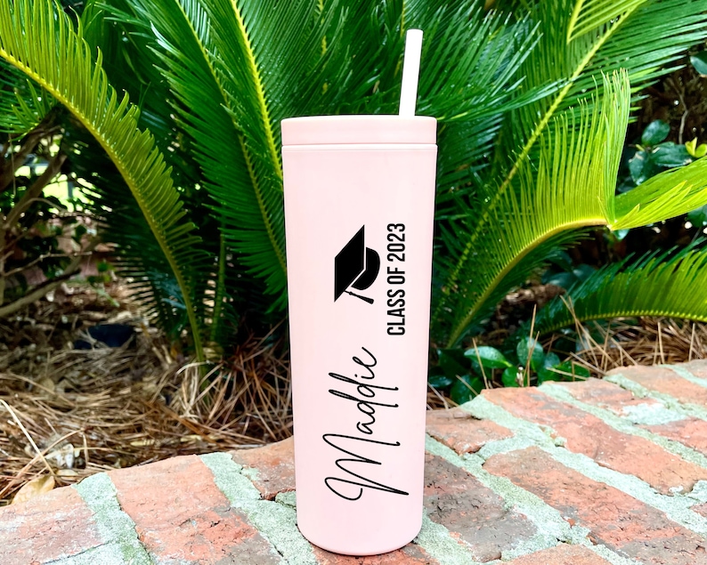 Puede incluir: Un vaso de acero inoxidable rosa con un gorro de graduaci&oacute;n negro y el texto "Class of 2023" y "Maddie" en letras negras.