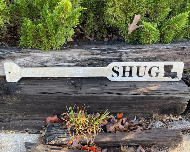 Puede incluir: Una pala de madera blanca con la palabra "SHUG" y un contorno del estado de Luisiana. La pala est&aacute; apoyada en un tronco de madera desgastado.