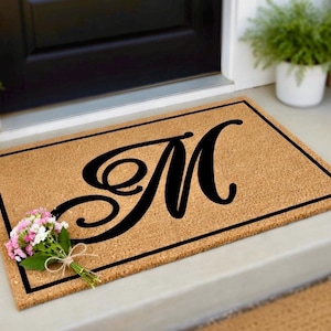 May include: A rectangular doormat with a black border and a large, elegant black letter "M" in the center. The mat is made of a natural, light brown material. A small bouquet of pink and white flowers rests on the mat.