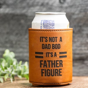 May include: A tan leather can cooler with the text "IT'S NOT A DAD BOD IT'S A FATHER FIGURE" in black. A white and silver can is inside the cooler. The background is a wooden surface.
