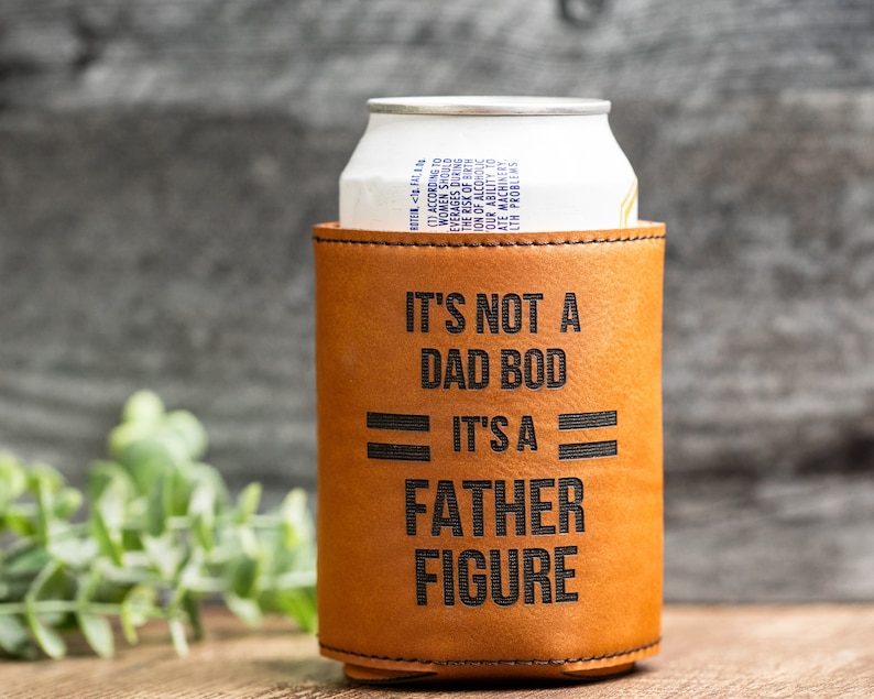 May include: A tan leather can cooler with the text "IT'S NOT A DAD BOD IT'S A FATHER FIGURE" in black. A white and silver can is inside the cooler. The background is a wooden surface.