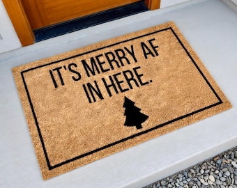 May include: A rectangular doormat with the text "IT'S MERRY AF IN HERE" in black, along with a black Christmas tree graphic. The mat is a natural brown color and has a black border.