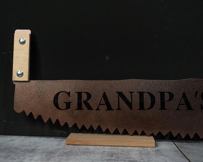 Puede incluir: Una hoja de sierra de metal r&uacute;stico con un mango de madera. La hoja est&aacute; pintada de color cobre y tiene la palabra "GRANDPA'S" recortada en negro.