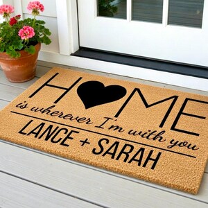 May include: A rectangular doormat with the word "HOME" in black letters, a black heart, and the phrase "is wherever I'm with you" below. The names "LANCE + SARAH" are printed at the bottom. The mat is tan and sits on a porch.