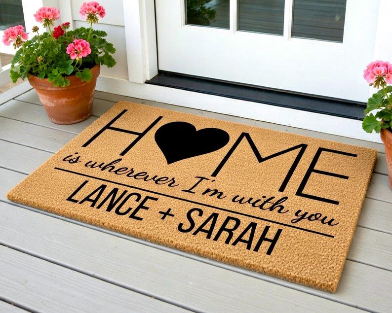 May include: A rectangular doormat with the word "HOME" in black letters, a black heart, and the phrase "is wherever I'm with you" below. The names "LANCE + SARAH" are printed at the bottom. The mat is tan and sits on a porch.