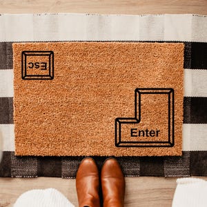 May include: A brown doormat with the words "ESC" and "Enter" printed on it in a black rectangular frame. The doormat is on a black and white striped rug.