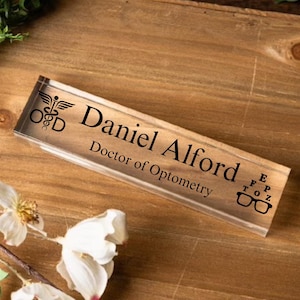 May include: A clear acrylic desk nameplate with black text that reads "Daniel Alford, Doctor of Optometry". The nameplate also features a black and white medical symbol and an eye chart with glasses.