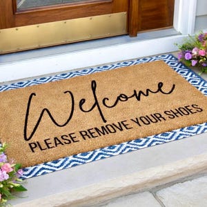 May include: A rectangular doormat with the word "Welcome" in a cursive font and the phrase "PLEASE REMOVE YOUR SHOES" in a block font. The mat is brown with a blue and white geometric border.