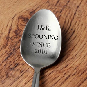 May include: A stainless steel spoon with the engraved text "J&K SPOONING SINCE 2010". The spoon is set against a wooden background, highlighting the inscription.