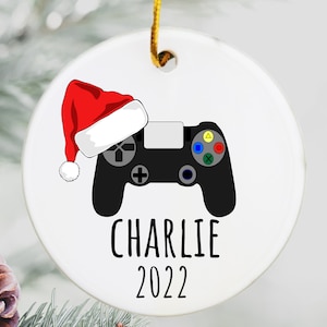 May include: White ceramic Christmas ornament with a black video game controller and a red Santa hat. The ornament has the name "CHARLIE" and the year "2022" printed on it.